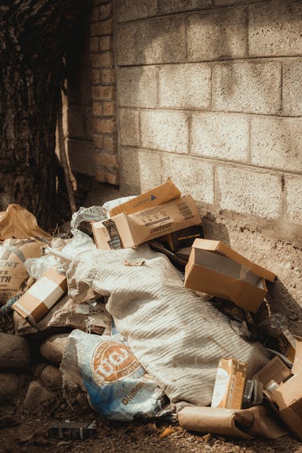 A pile of discarded cardboard boxes, some partially flattened and others still in their rectangular shape, rests against a weathered brick wall outdoors. The boxes vary in size and are made of brown k