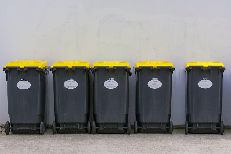 Five large plastic wheeled rubbish bins with black bodies and bright yellow lids are lined up side by side against a plain, off-white wall in an outdoor setting. The bins are arranged on a smooth conc