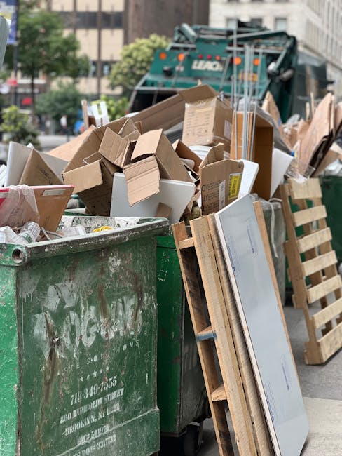 Five large plastic wheeled rubbish bins with black bodies and bright yellow lids are lined up side by side against a plain, off-white wall in an outdoor setting. The bins are arranged on a smooth conc