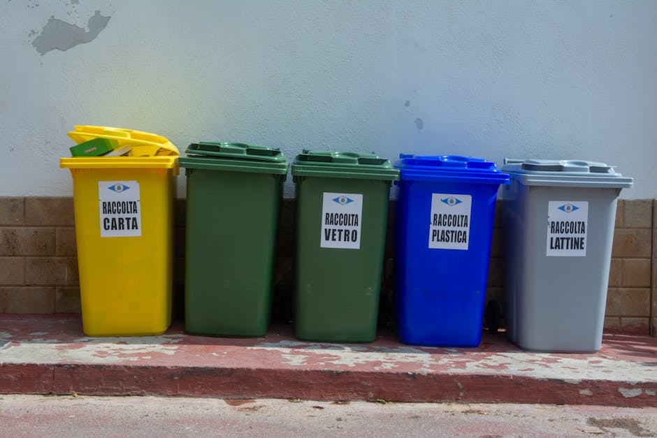Four individual waste bins aligned on a paved area adjacent to a garden or park, each designed for separate recycling purposes. The first bin from the left is light blue with a textured surface, inten